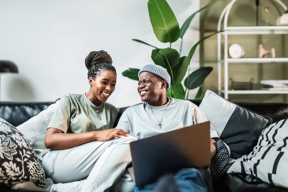 A happy young Black couple laughing together while relaxing on a cozy sofa with a laptop.