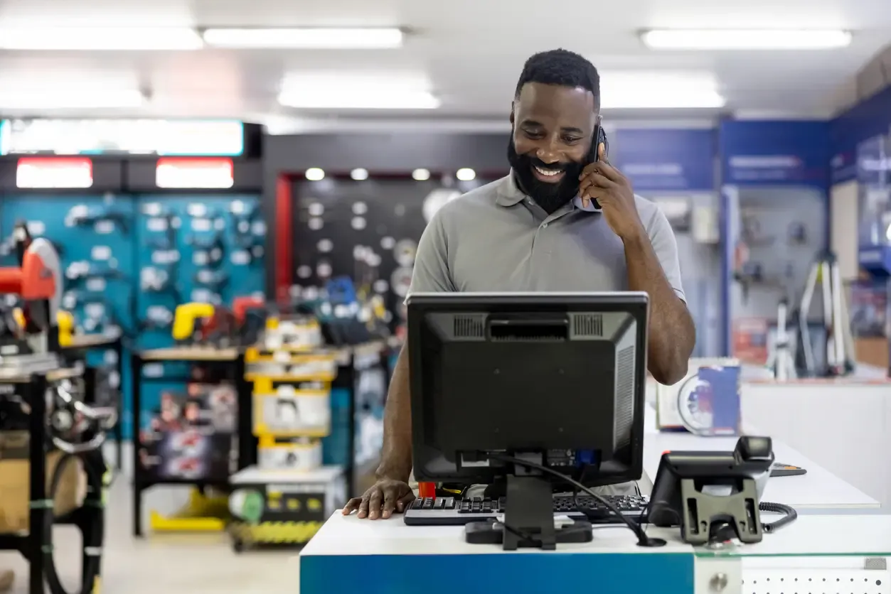 A smiling male employee in a grey polo shirt stands behind a service counter at a hardware store while talking on a smartphone and looking at a computer monitor.