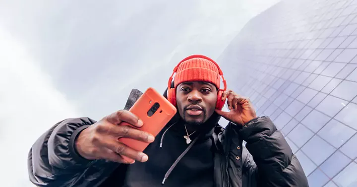 Man with orange phone, headphones and beanie, looking at his phone.