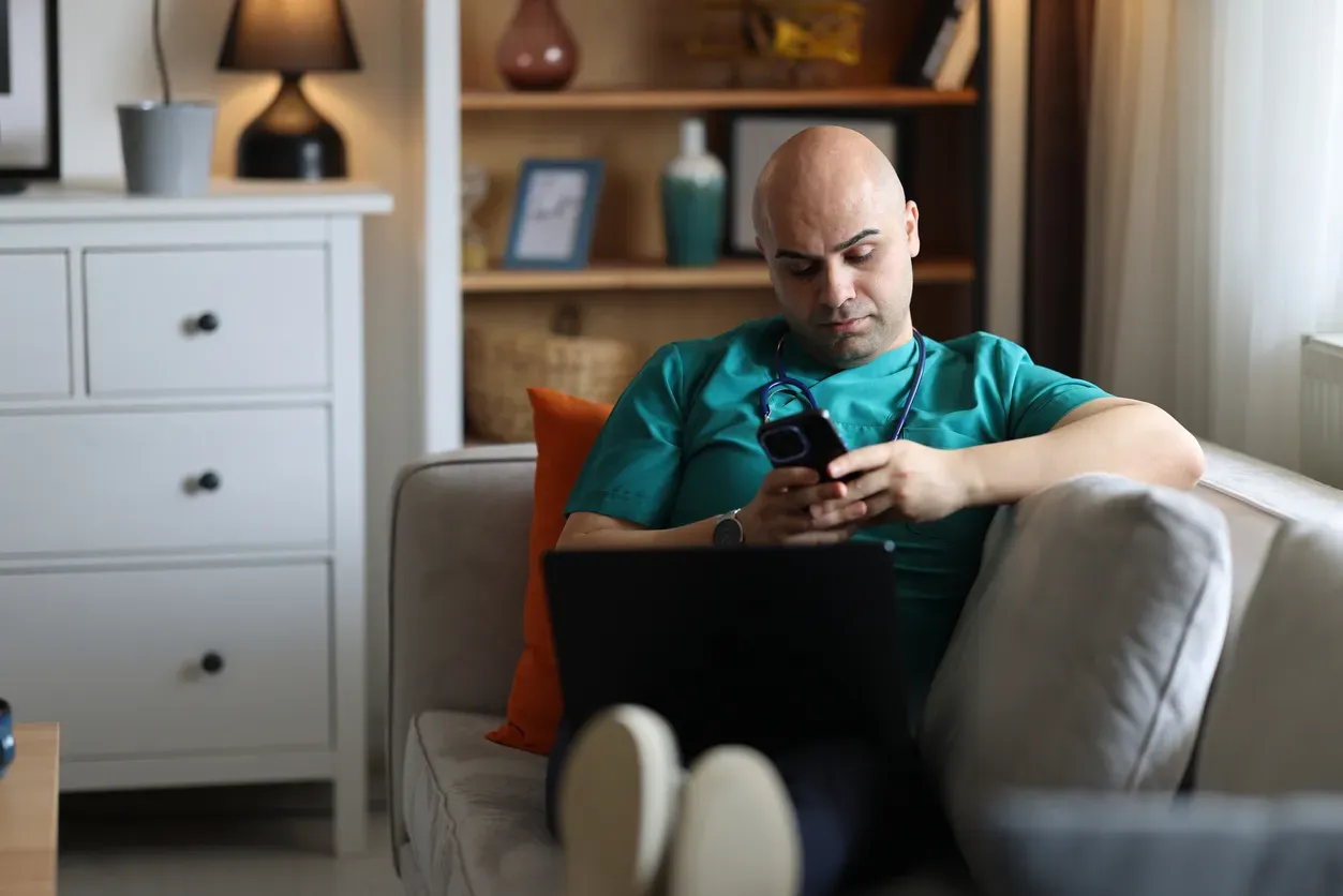 A man in teal scrubs and a stethoscope checks his phone on a sofa.