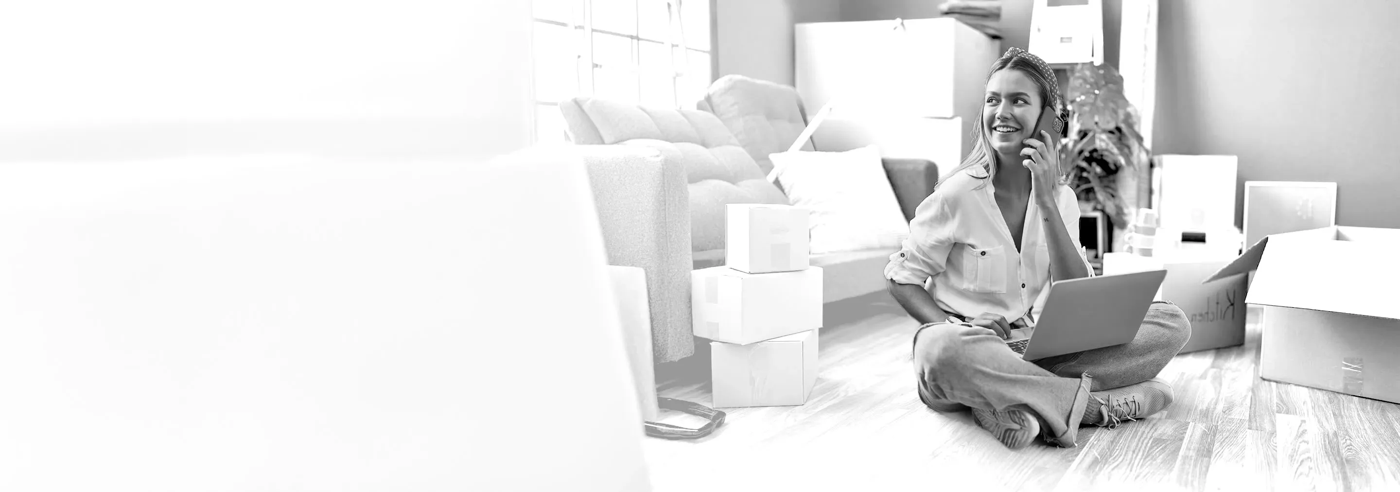 A smiling woman sits on the floor of a new home surrounded by moving boxes, using a laptop and talking on a smartphone.