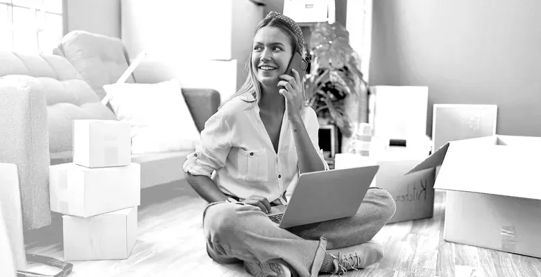 A smiling woman sits on the floor of a new home surrounded by moving boxes, using a laptop and talking on a smartphone.