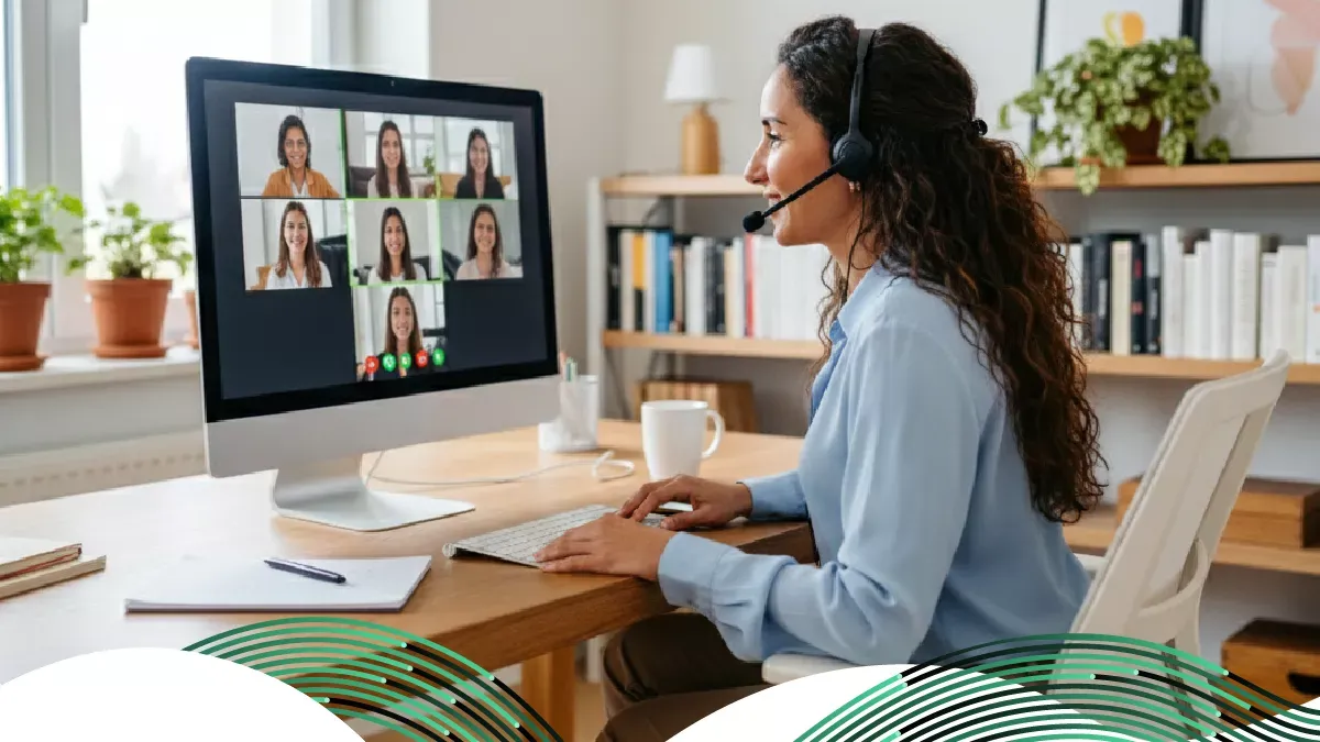 Una mujer con auriculares se sienta en un escritorio en una oficina en casa iluminada mientras participa en una videoconferencia en un monitor grande.
