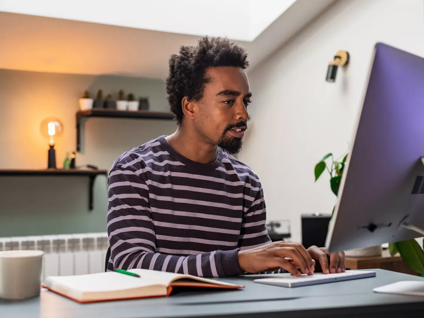 A focused man in a striped shirt types on a computer keyboard at his desk in a well-lit home office.