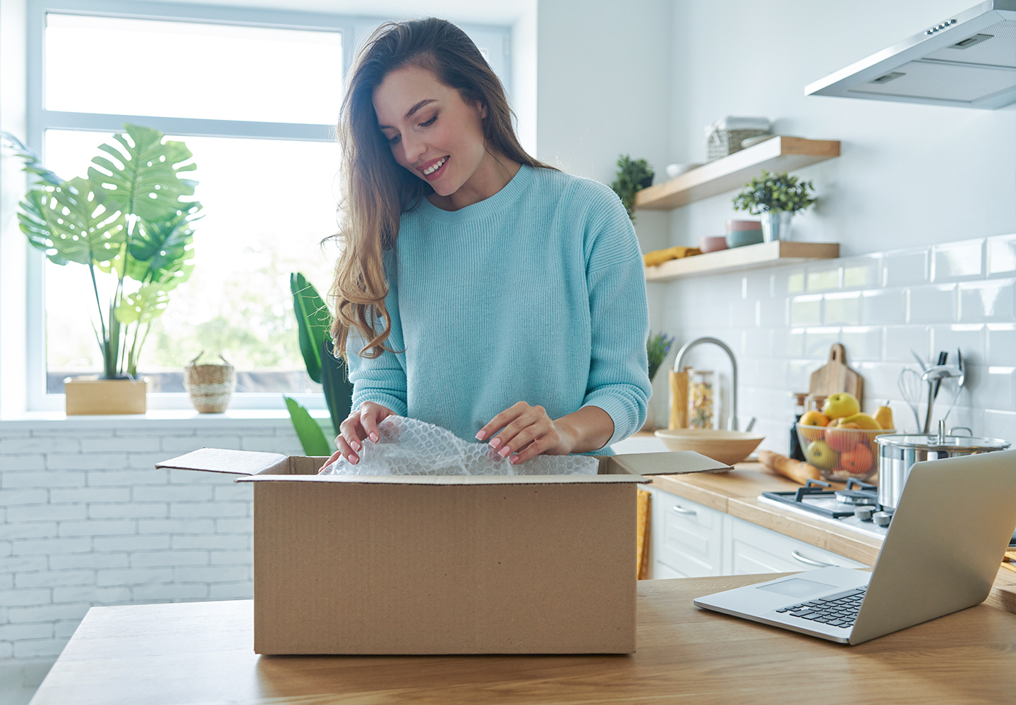 Woman in kitchen with laptop, opening a package