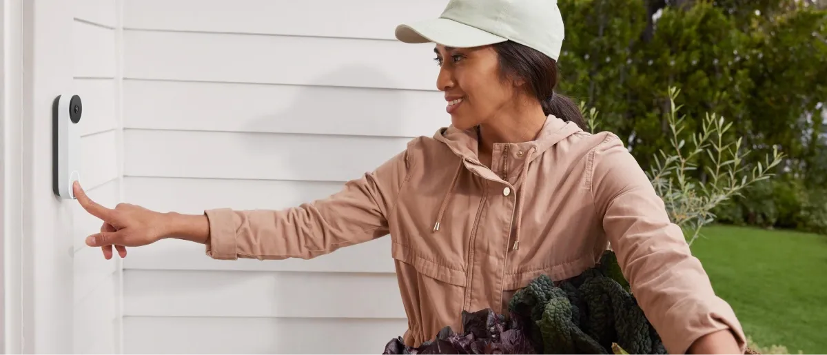 A woman holding groceries presses a smart video doorbell on a house.