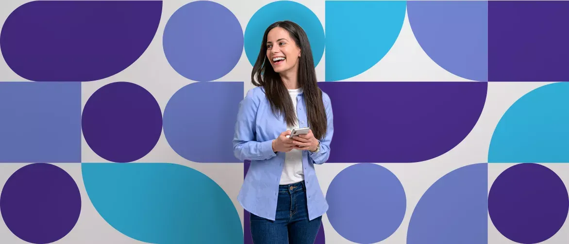 Generate alt text for this image A happy woman in a blue shirt holding a phone, standing in front of a blue and purple geometric background.