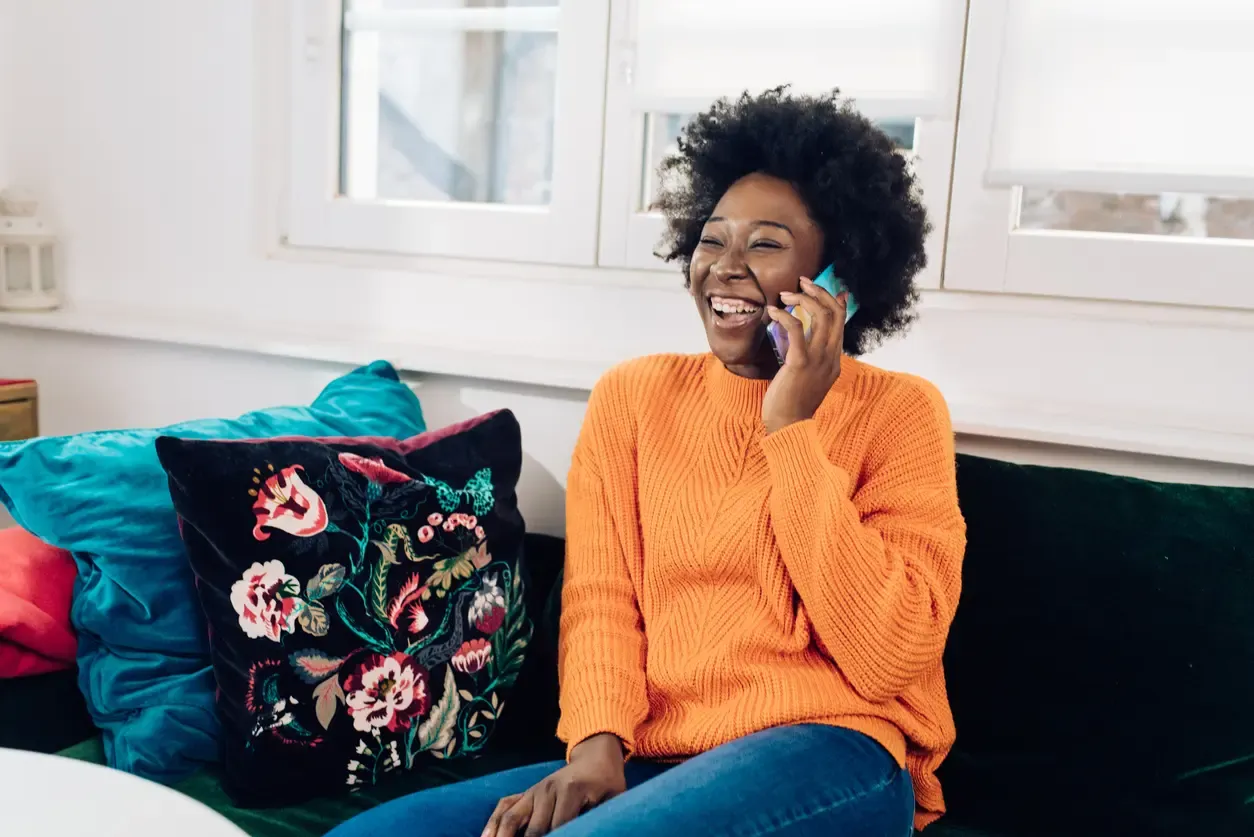 A laughing Black woman with an afro wearing an orange sweater and sitting on a sofa while talking on a smartphone.