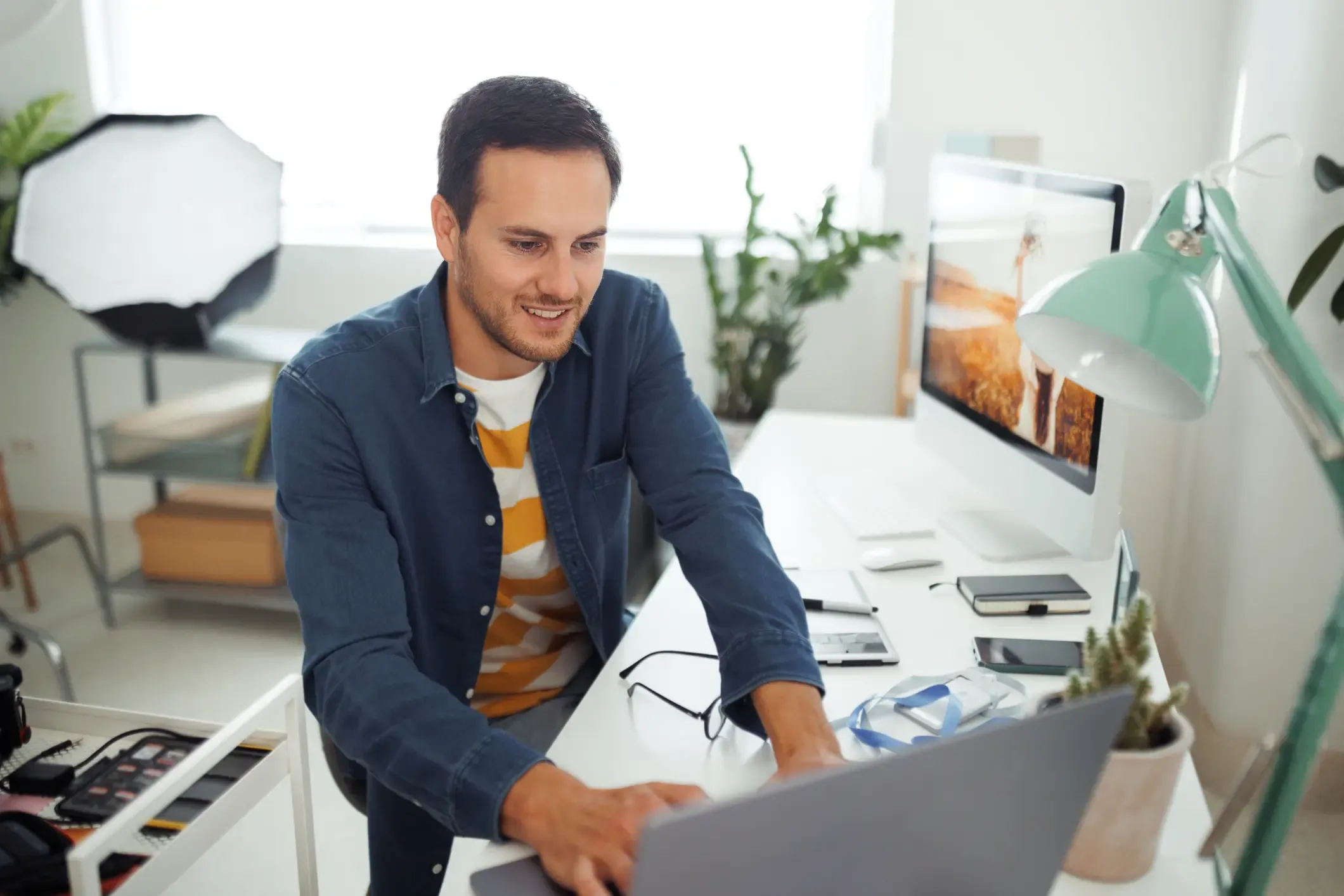 A smiling man in a blue shirt sitting at a white desk and working on a laptop in a bright, modern home office or studio.