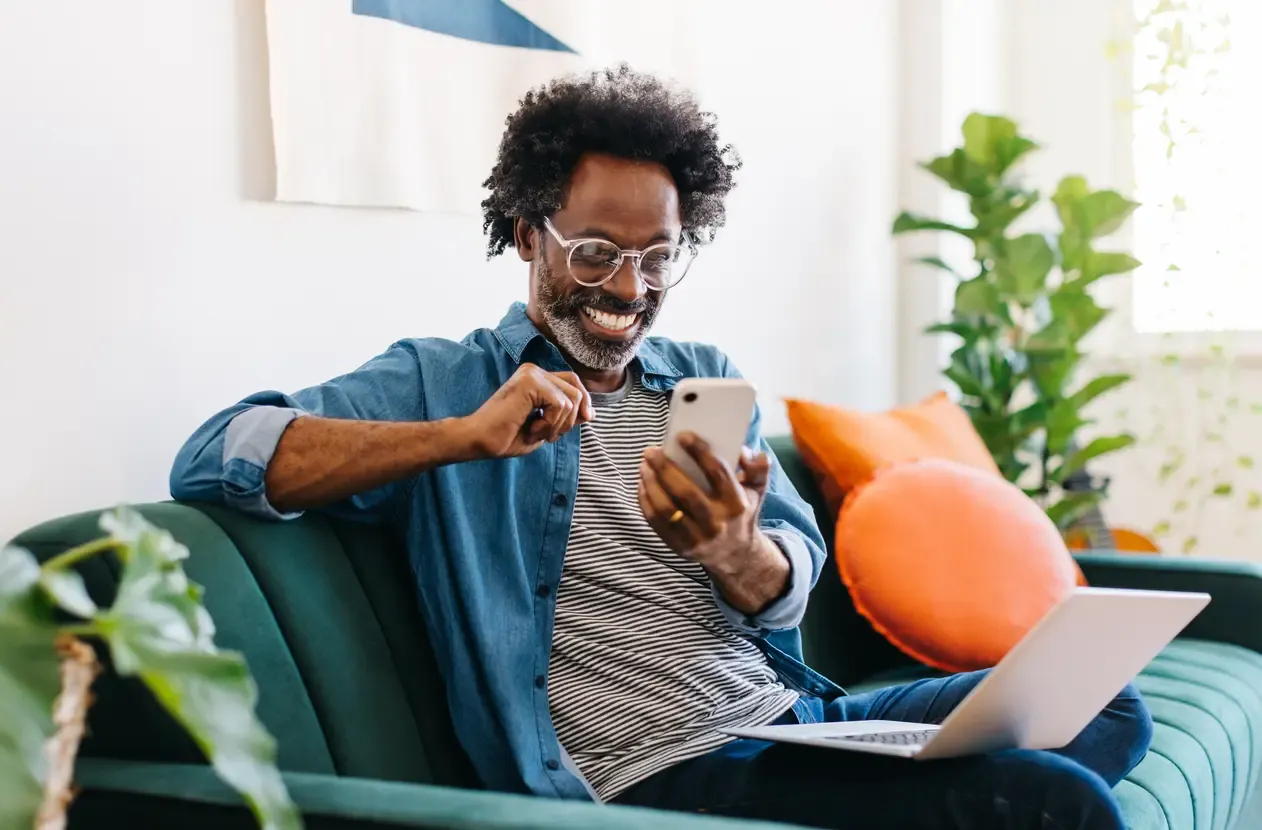 A smiling man pumps his fist while looking at his phone with a laptop nearby.