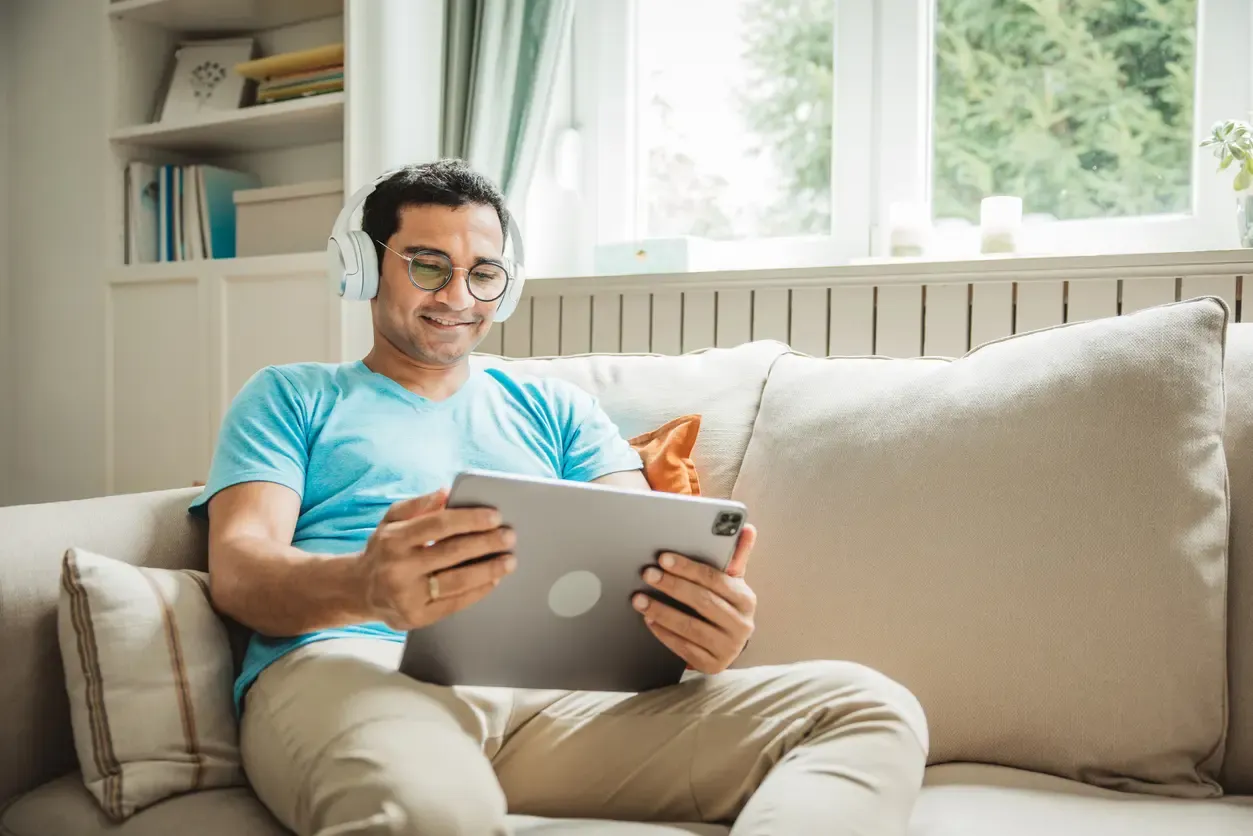 A smiling man wearing white headphones watches his tablet while relaxing on a couch.