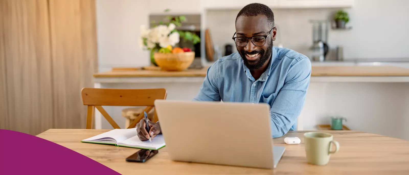 A man with glasses smiling while using a laptop and taking notes at a kitchen table.