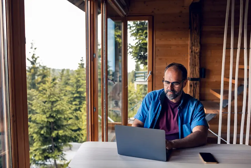 A middle-aged man with glasses working on a laptop at a table inside a wooden cabin with a view of evergreen trees.