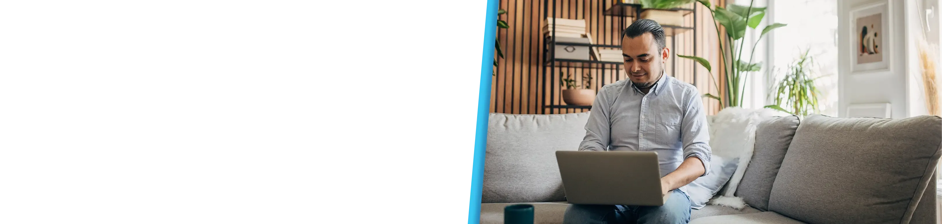 A man in a grey shirt works on a laptop while sitting on a grey couch at home.