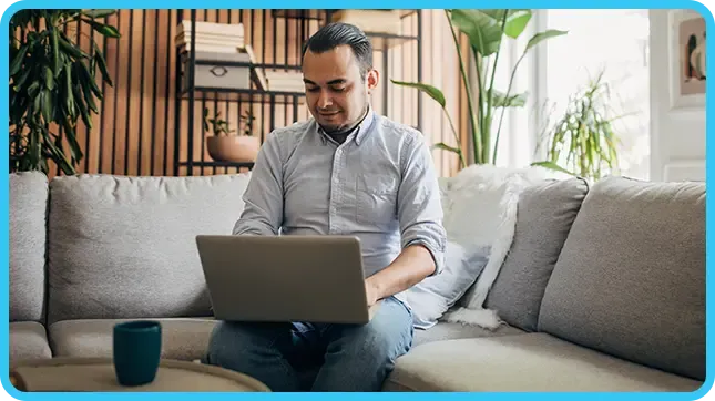 A man in a grey shirt works on a laptop while sitting on a grey couch at home.