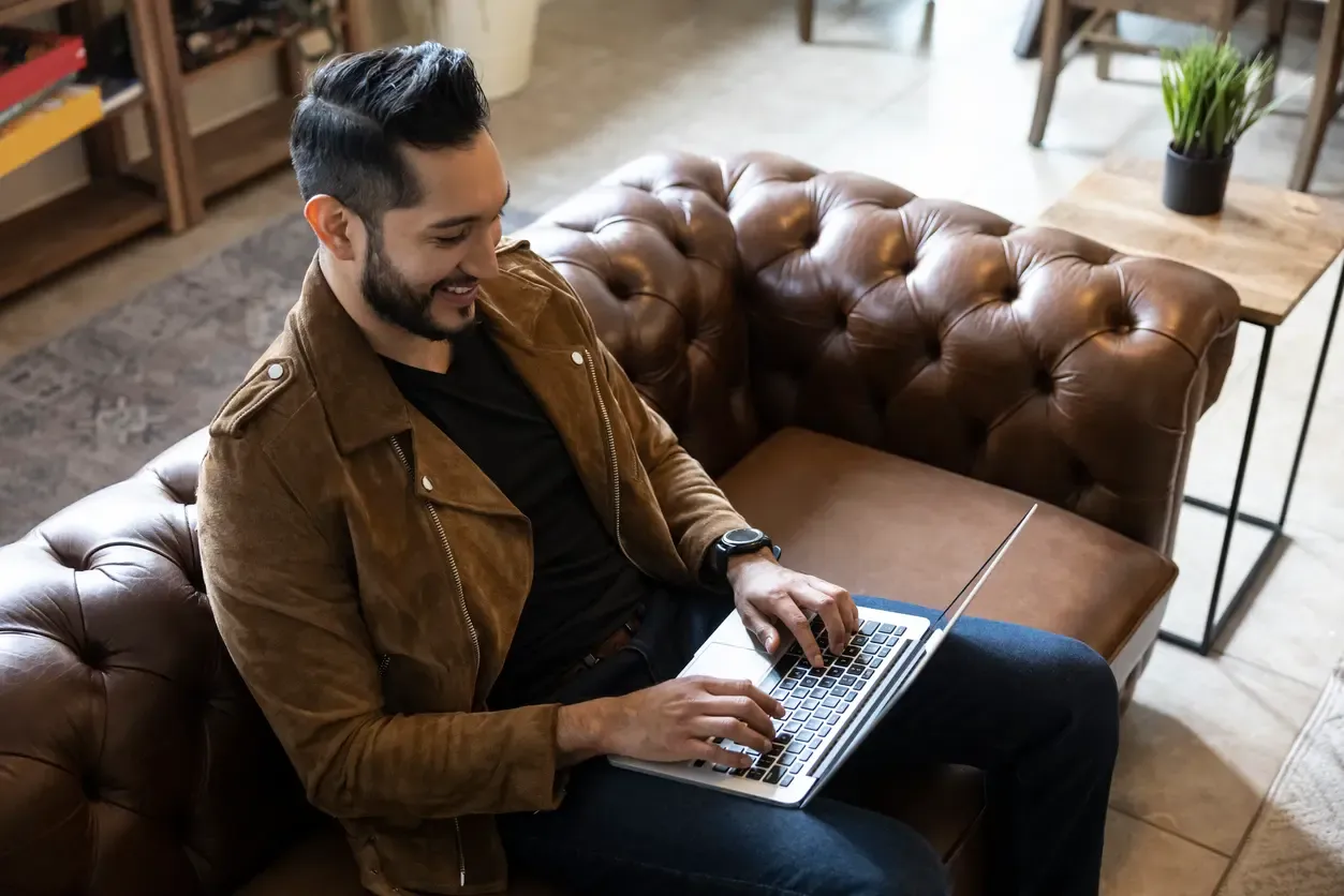 A smiling man in a brown jacket types on a laptop while seated on a tufted leather sofa.