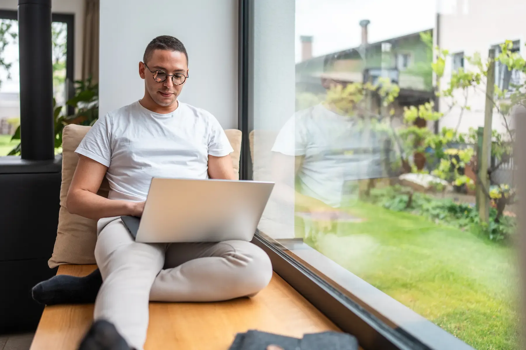 A man in glasses works on his laptop while sitting on a cushioned bench by a large window.
