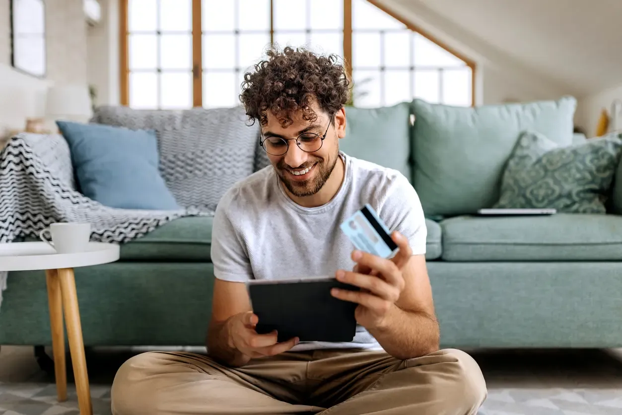 A smiling man sits on a living room floor holding a tablet and credit card while shopping online