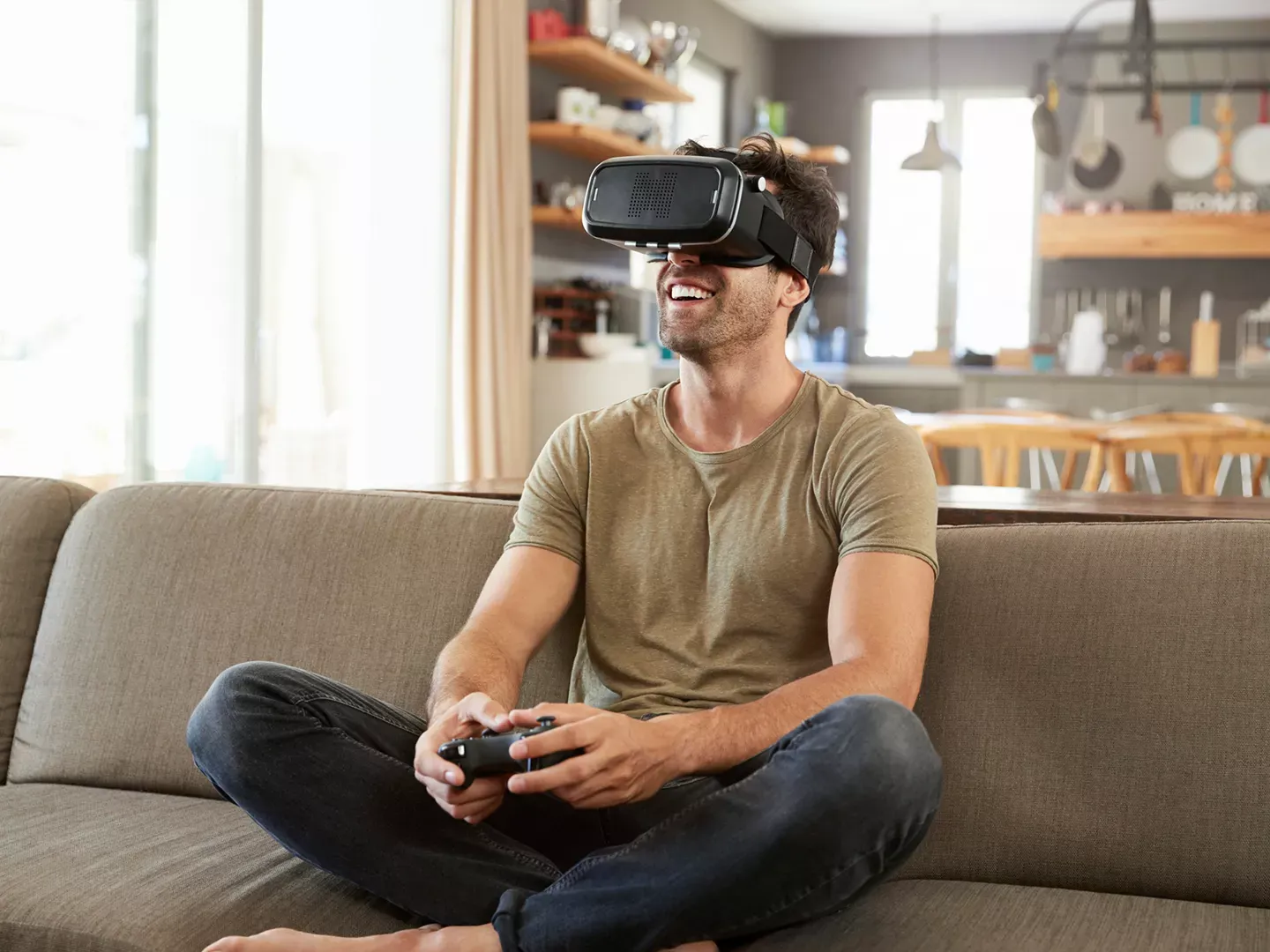 A man smiling and wearing a virtual reality headset while sitting cross-legged on a couch and holding a gaming controller.