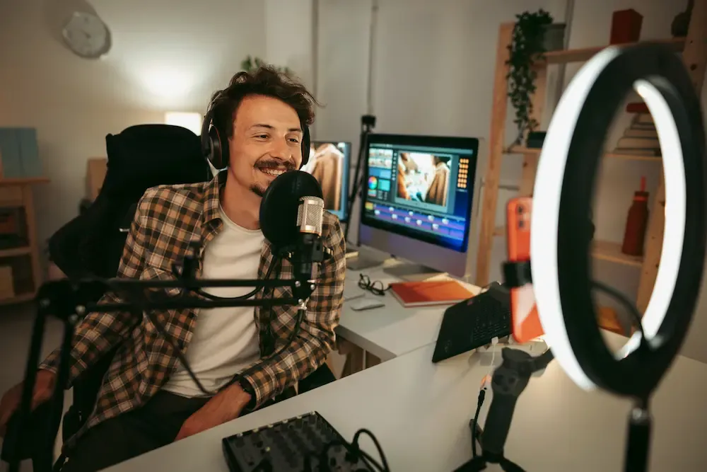 A smiling man wearing headphones speaks into a professional microphone while recording a video or podcast in a home studio with a ring light and computer monitors.