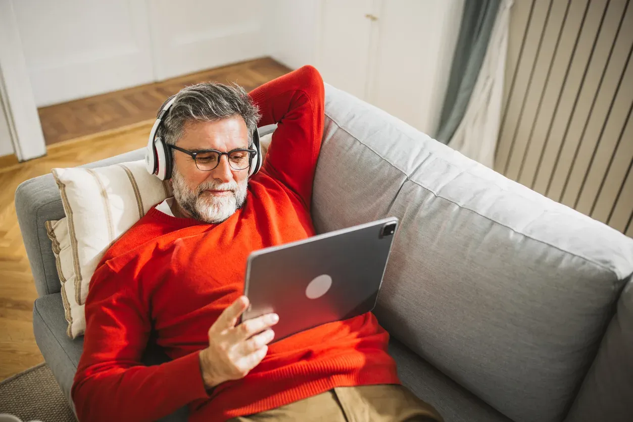 A man in a red sweater relaxes on a couch using a tablet with headphones on.