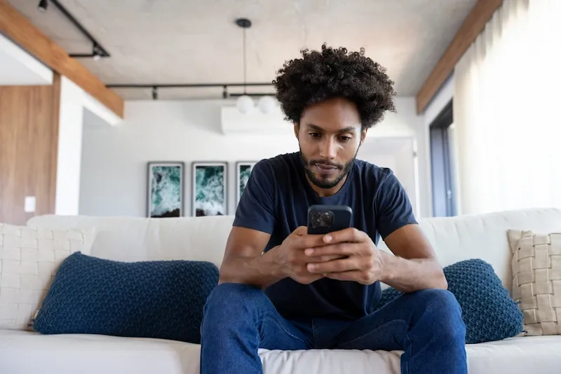 A man sits on a white sofa in a modern living room looking at his smartphone.