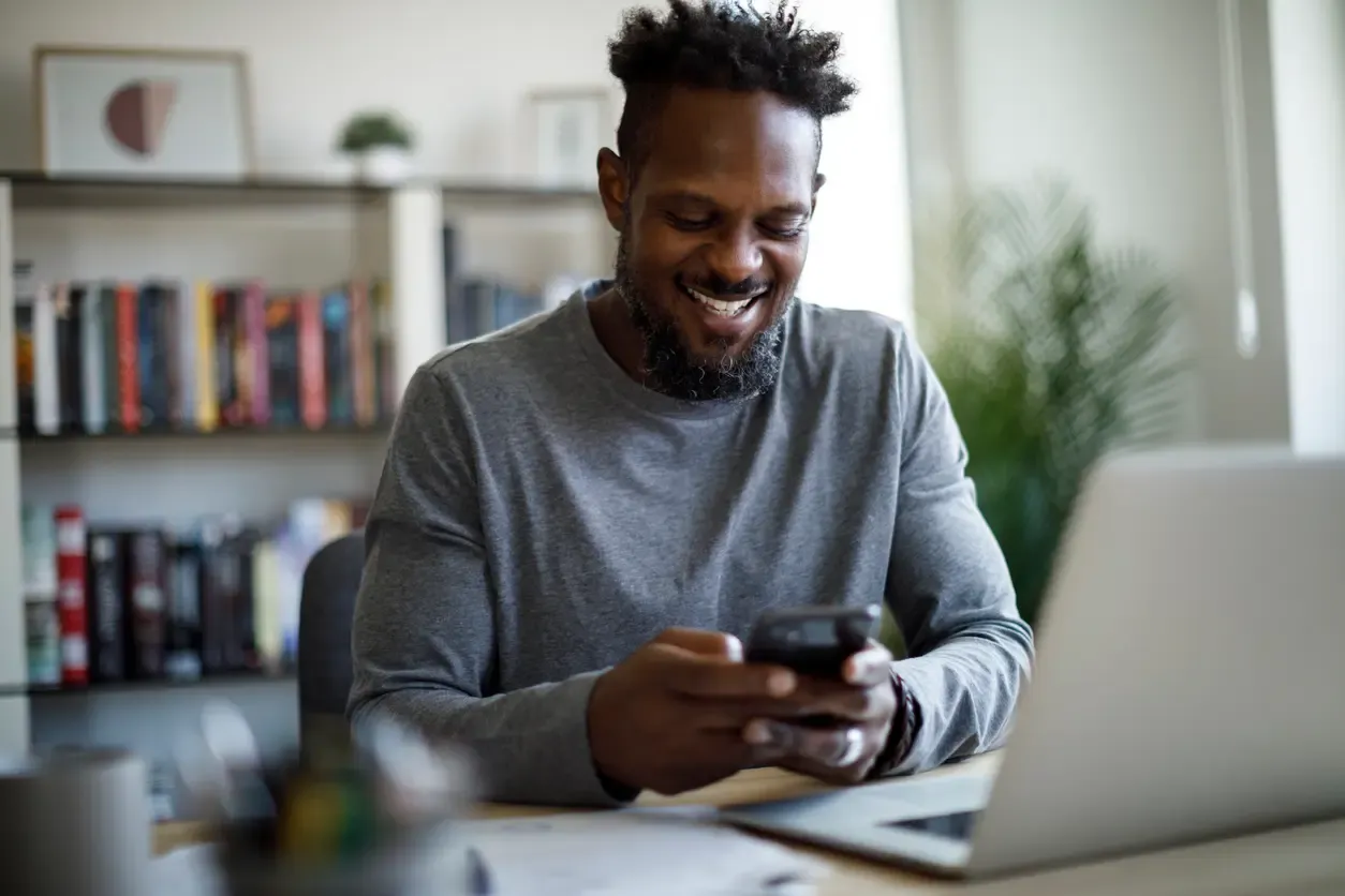 A smiling man uses his smartphone at a home office desk beside a laptop.