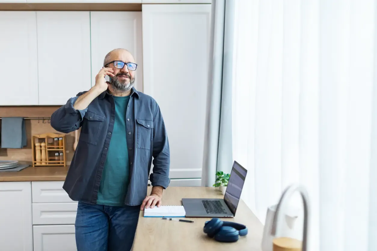 A middle-aged man with glasses talking on a smartphone while standing at a kitchen counter used as a workspace with a laptop and notebook.
