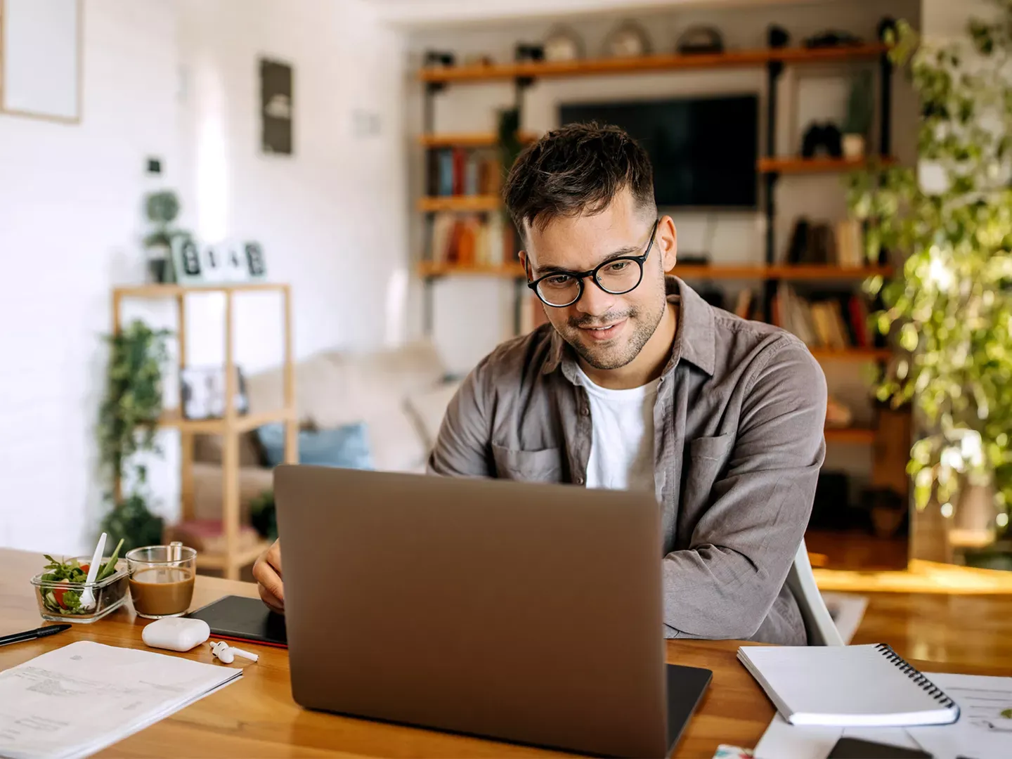 A man wearing glasses works on a laptop at a wooden desk in a bright home office.