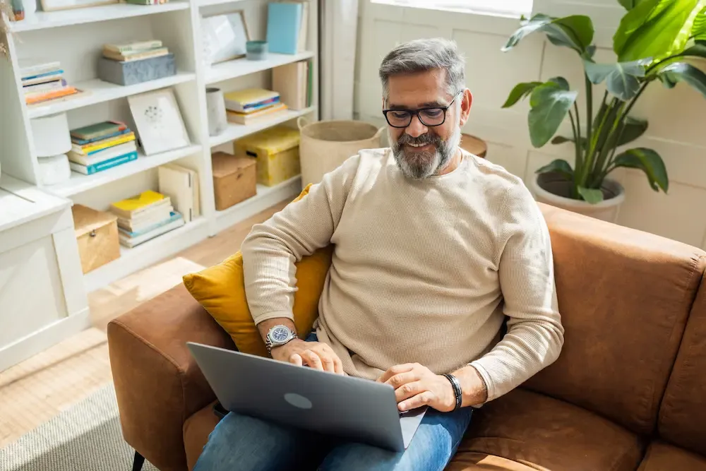 A smiling middle-aged man with glasses sitting on a brown leather couch and using a laptop in a bright living room.
