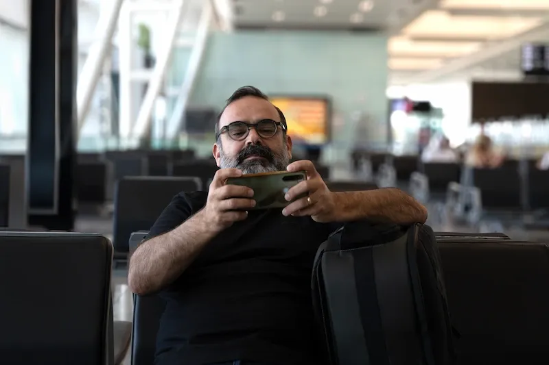 A bearded man with glasses sits in an airport waiting area, holding his smartphone horizontally with both hands.