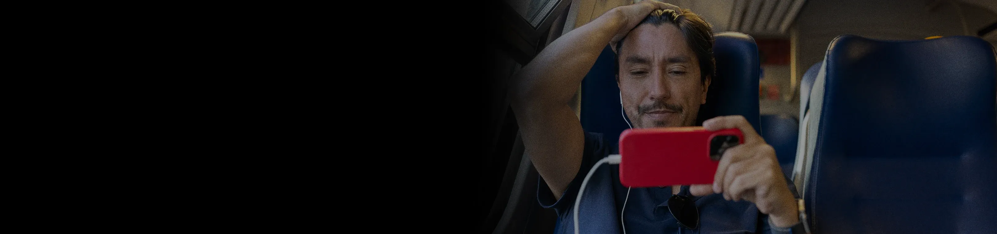 A man sitting on a train looks at his red smartphone with wired headphones, resting one hand on his head.