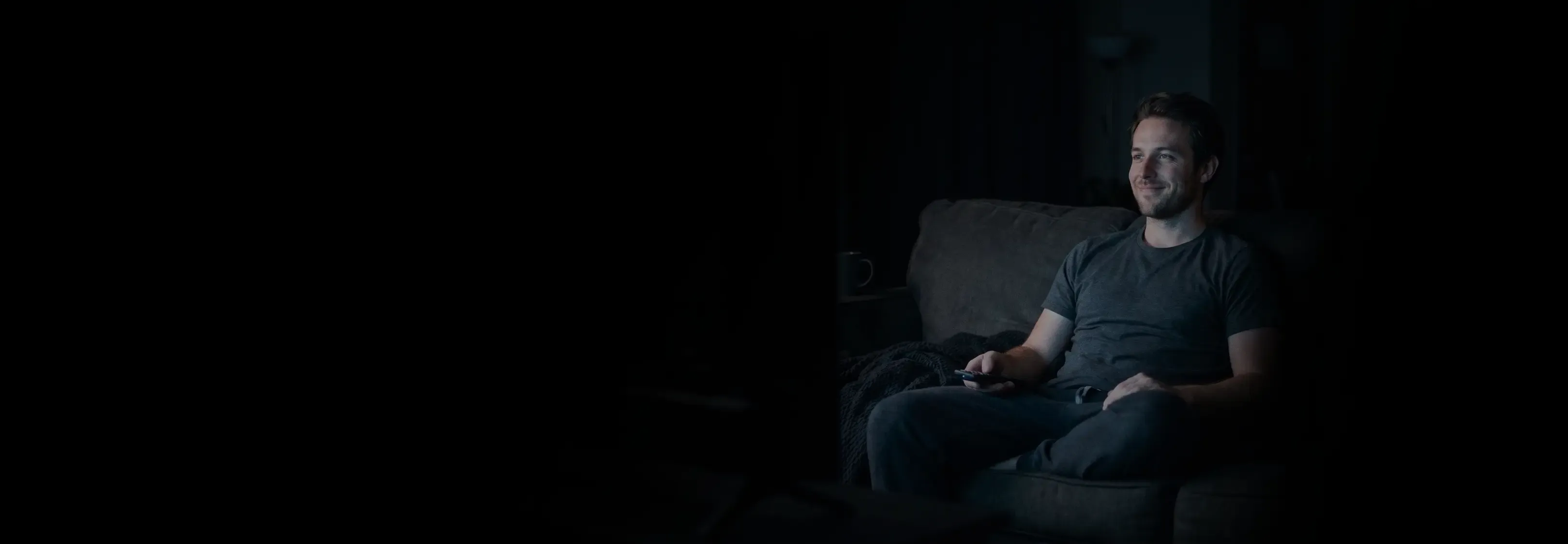 A smiling man holds a remote while relaxing on a gray sofa in a darkened living room lit by a TV screen.