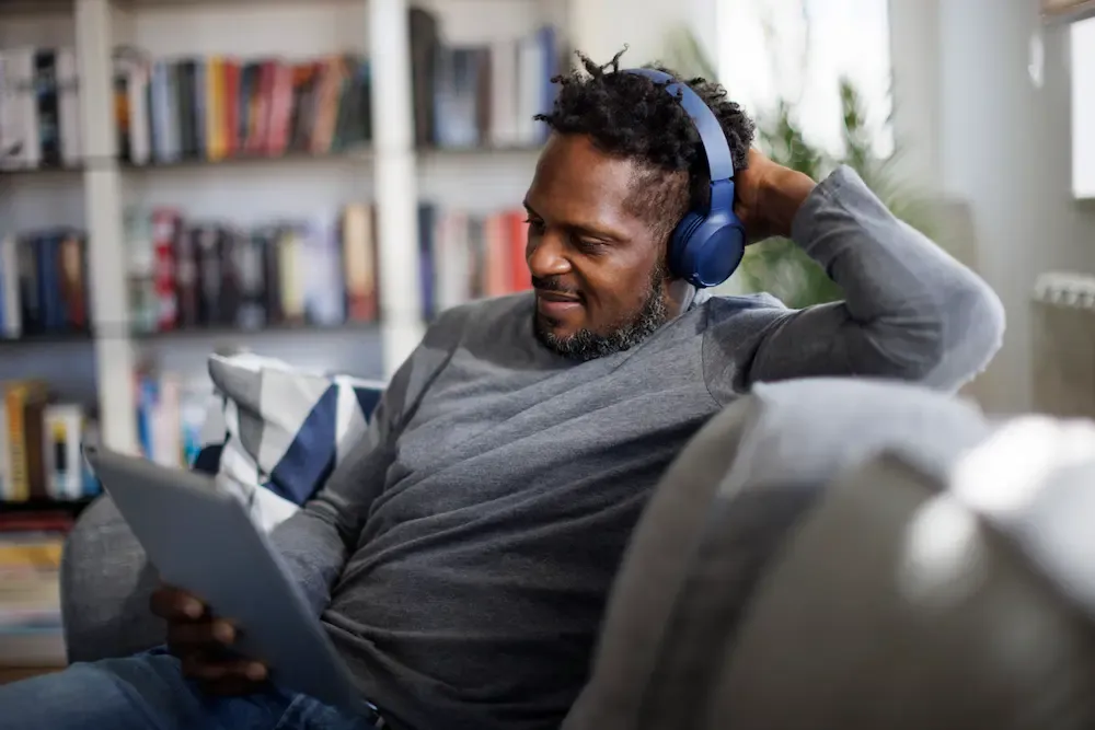 A man wearing blue headphones relaxes on a sofa while looking at a tablet in a room with bookshelves.