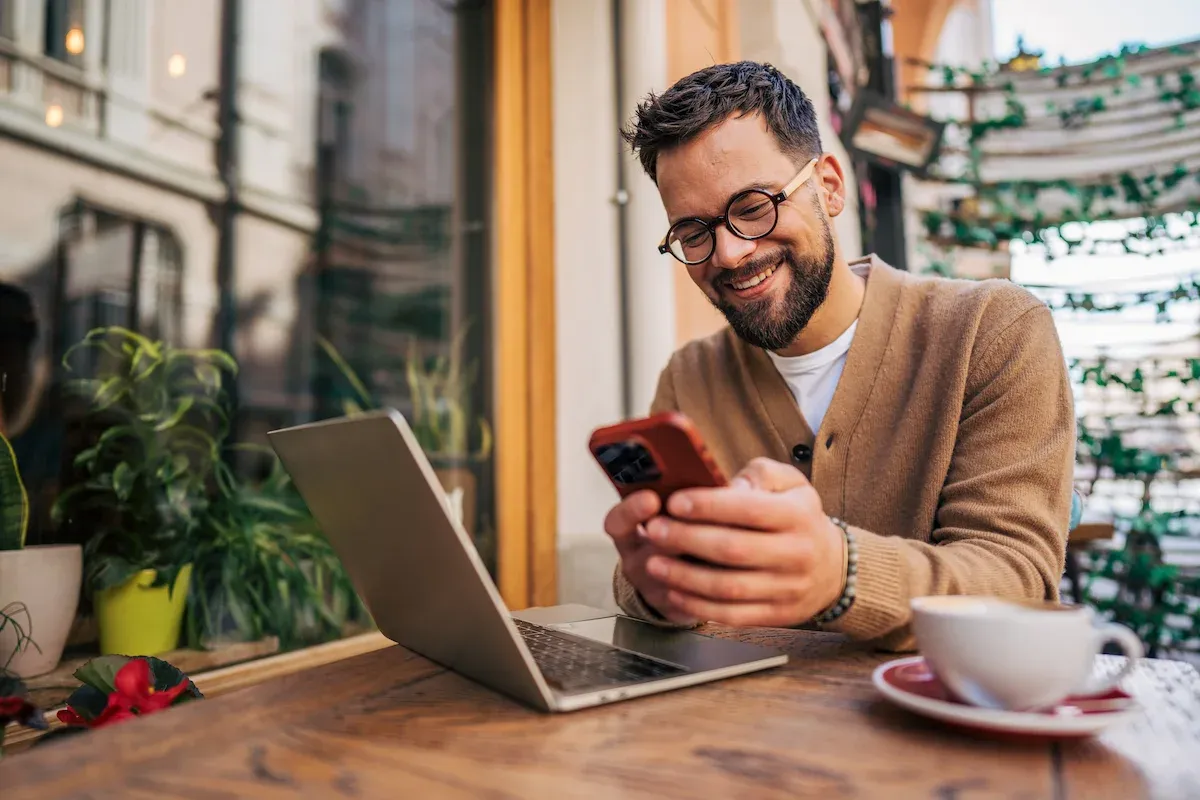 A bearded man in glasses smiles at his phone beside a laptop at a café.