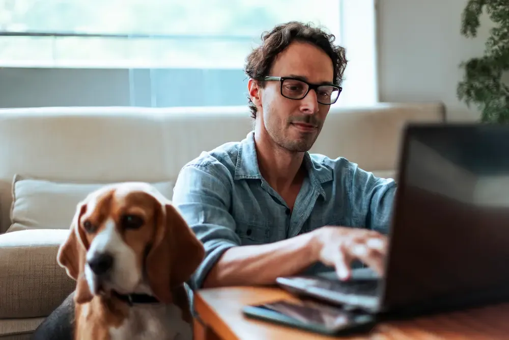 A man wearing glasses sitting on the floor and working on a laptop while his beagle dog sits beside him.