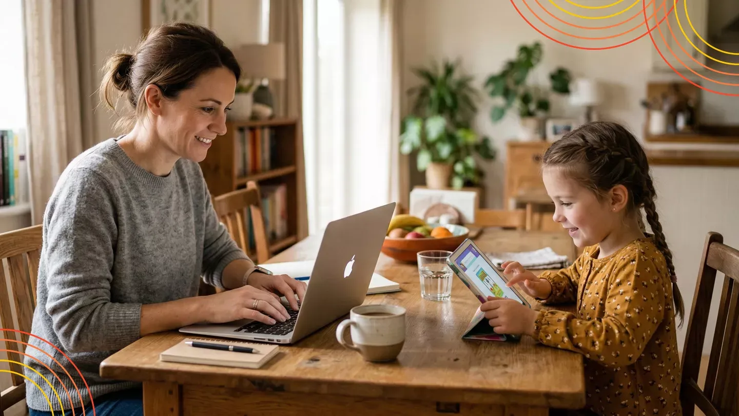 A woman typing on a laptop while a young girl sits next to her playing on a tablet at a wooden dining table.