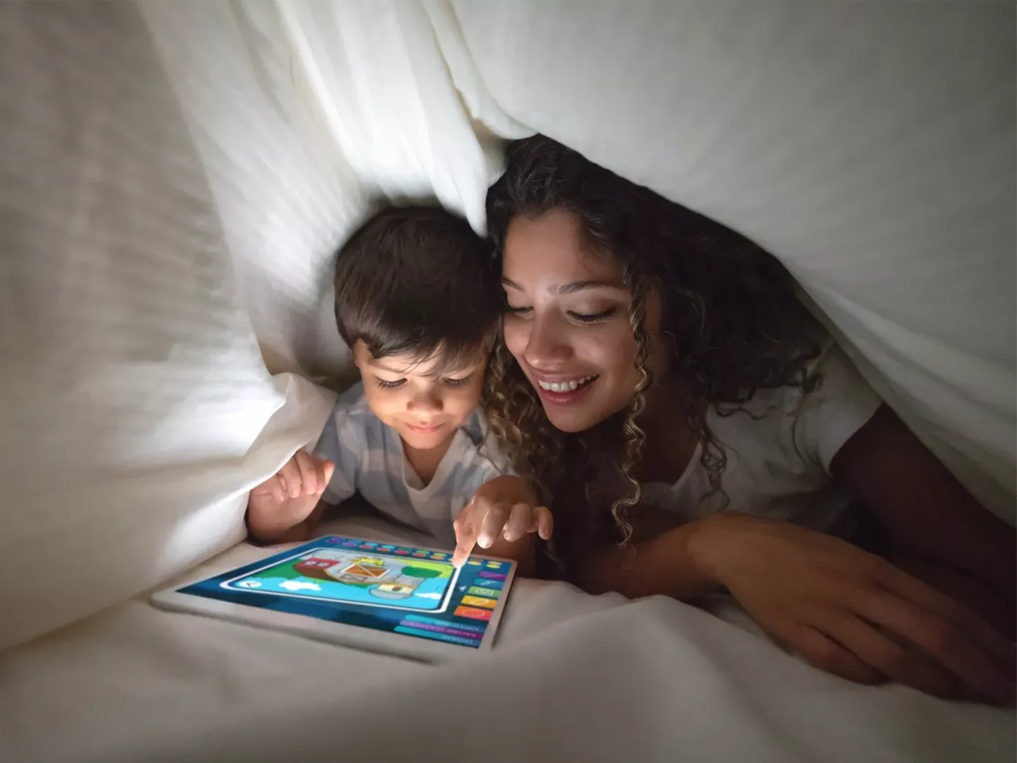 A mother and her young son smiling while playing an educational game on a tablet inside a cozy blanket fort.