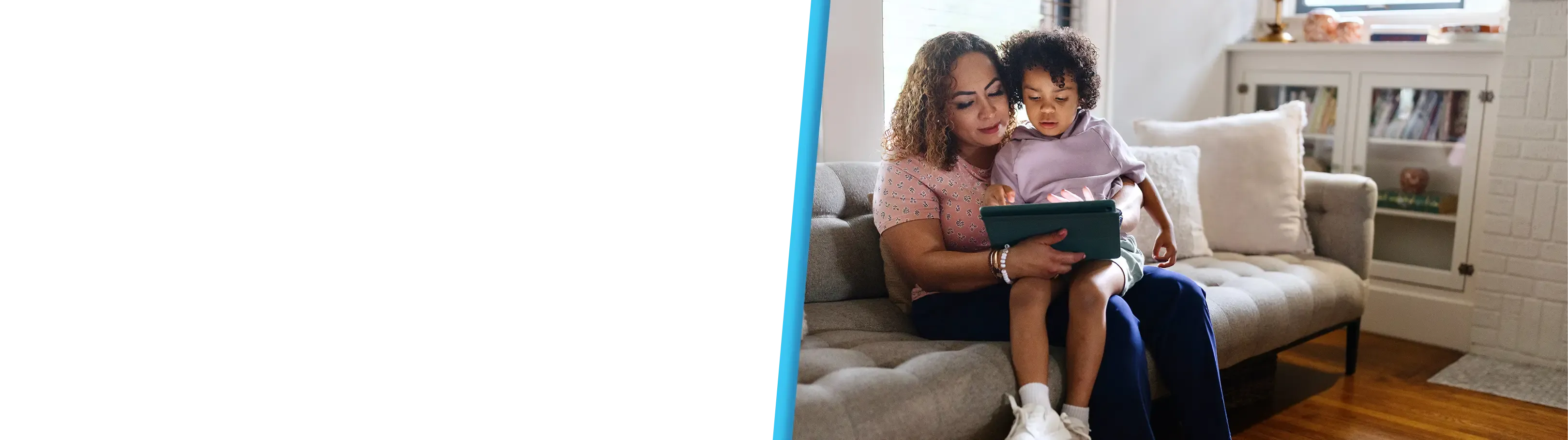 A mother sits on a grey sofa with her young child on her lap as they both look at and interact with a digital tablet together.