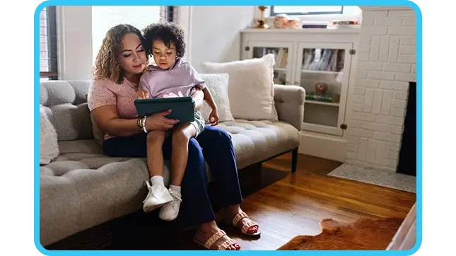 A mother sits on a grey sofa with her young child on her lap as they both look at and interact with a digital tablet together.