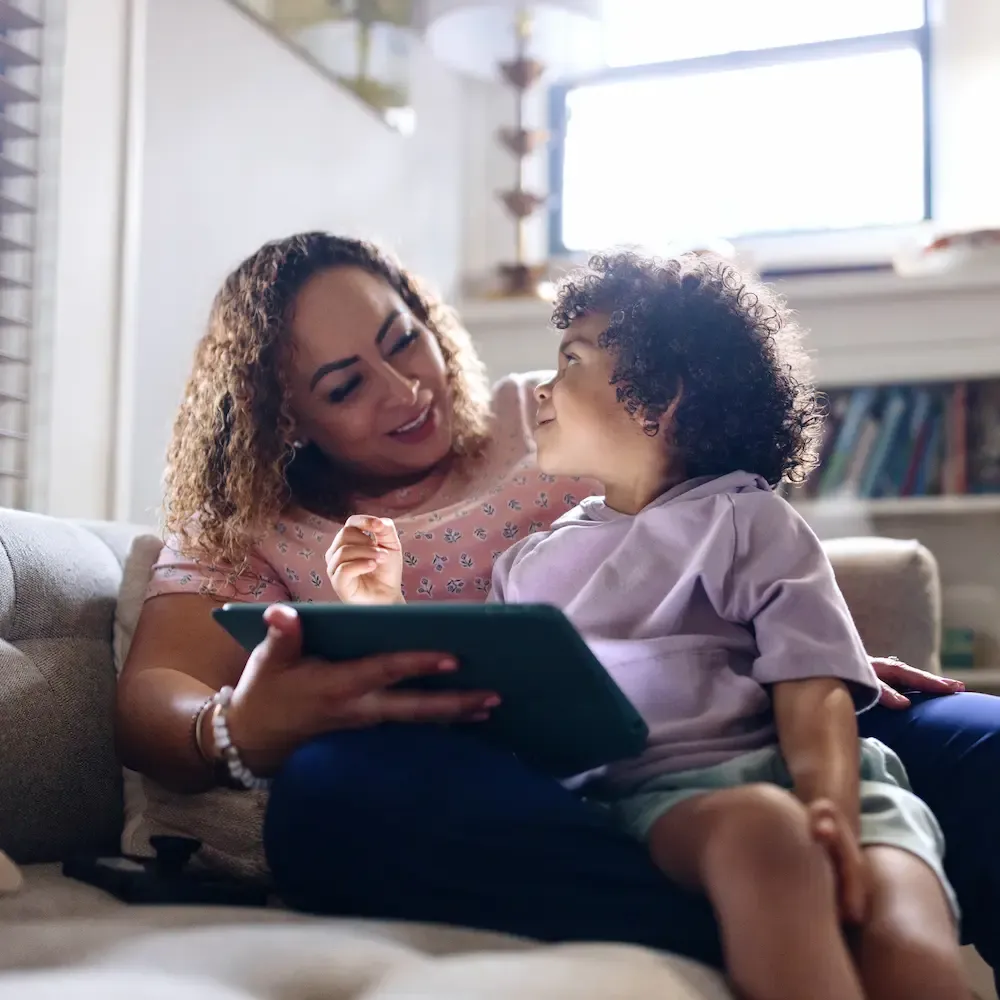 A smiling mother sits on a sofa holding a tablet while her young child looks up at her.