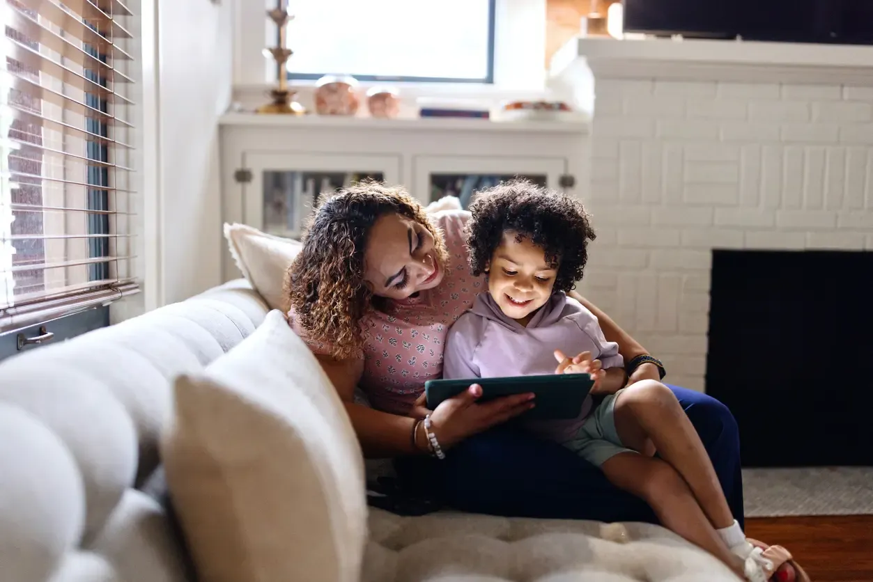 A mother and her young child smile while looking at a tablet together on the living room couch.