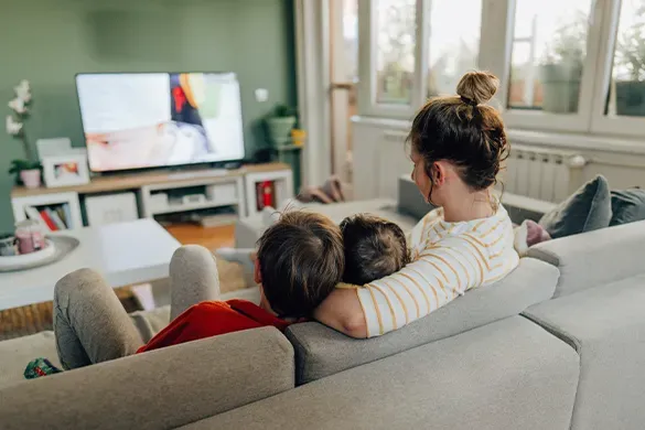 A mother cuddles two children on a sofa while they watch television together.