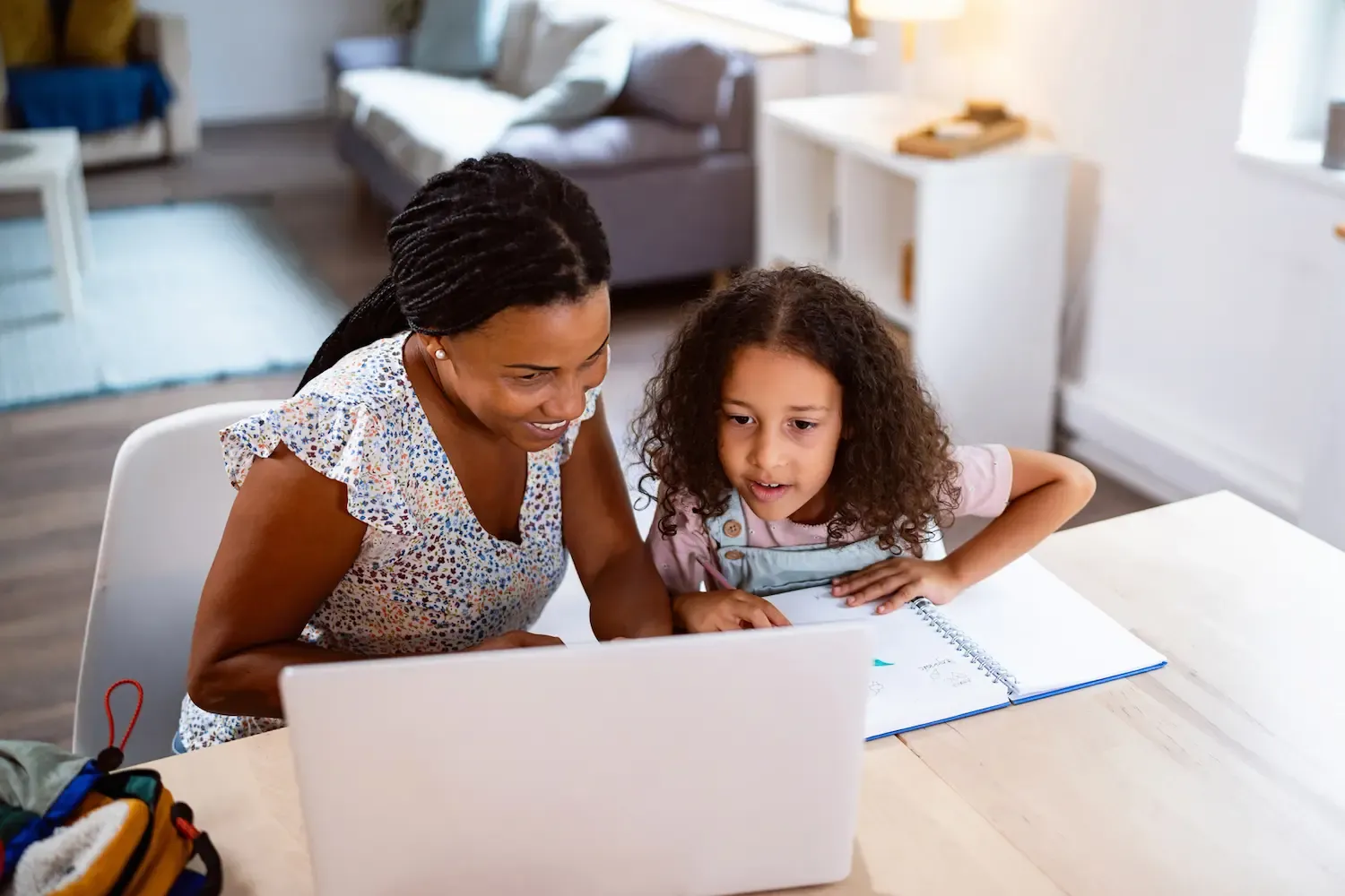 A woman and a young girl sit at a wooden table together looking at a laptop and a spiral notebook in a bright living room.