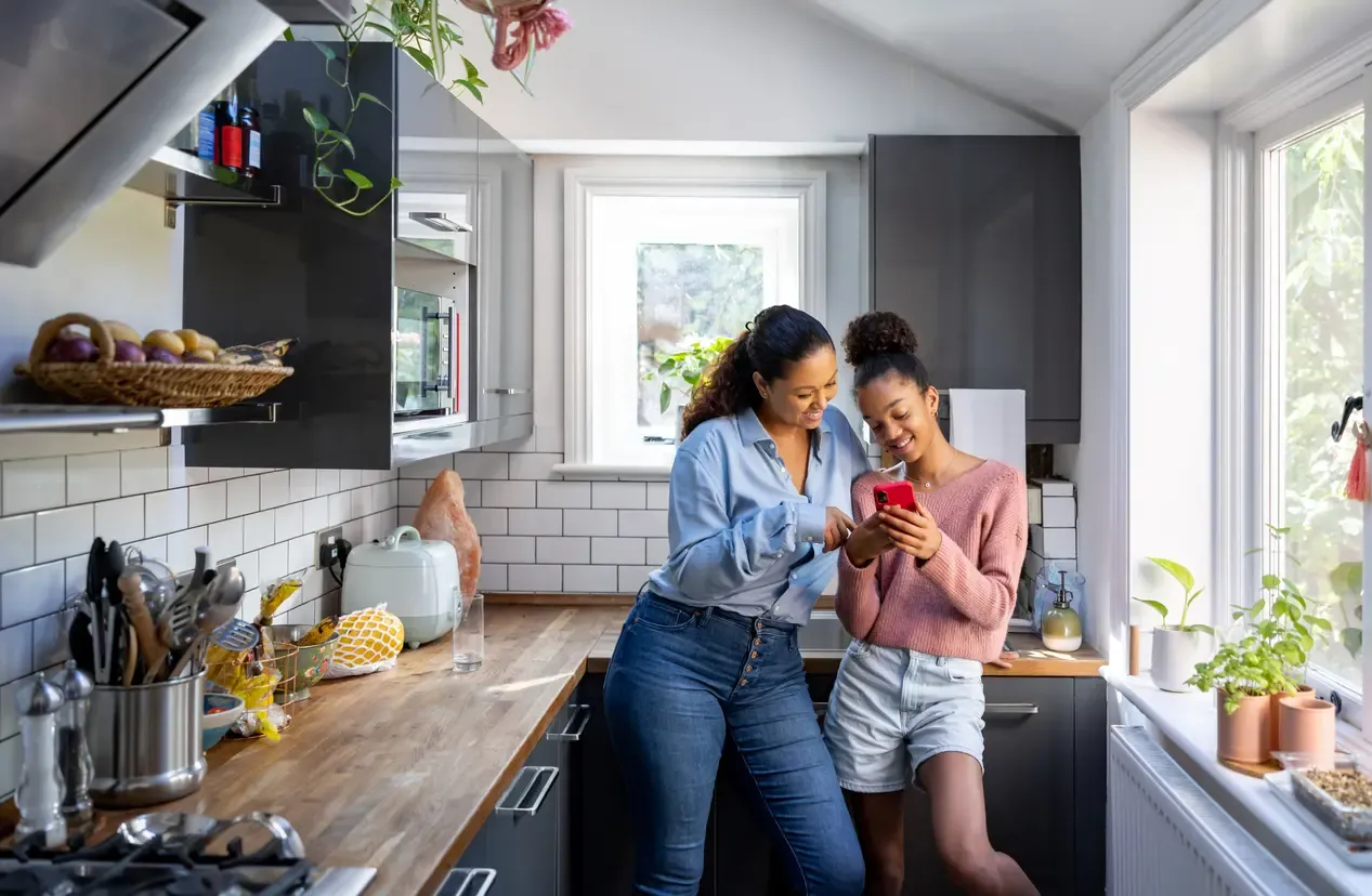 A mother and teenage daughter smile while looking at a smartphone together in their kitchen.