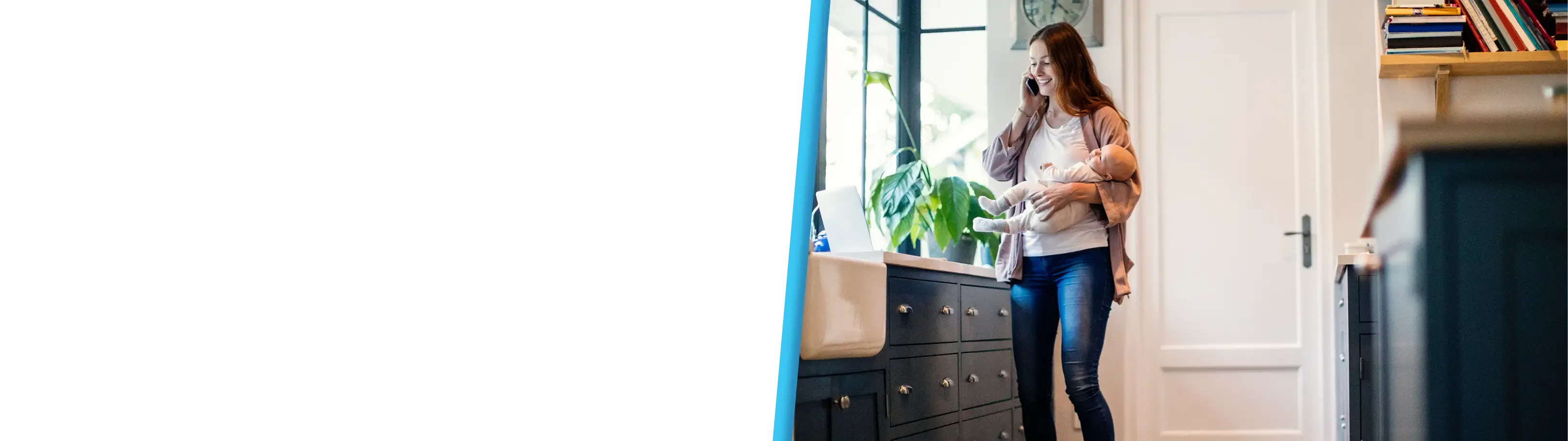 A smiling woman talks on the phone while holding a baby in a dark-cabineted kitchen.