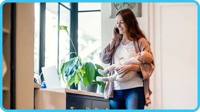 A smiling woman talks on the phone while holding a baby in a dark-cabineted kitchen.