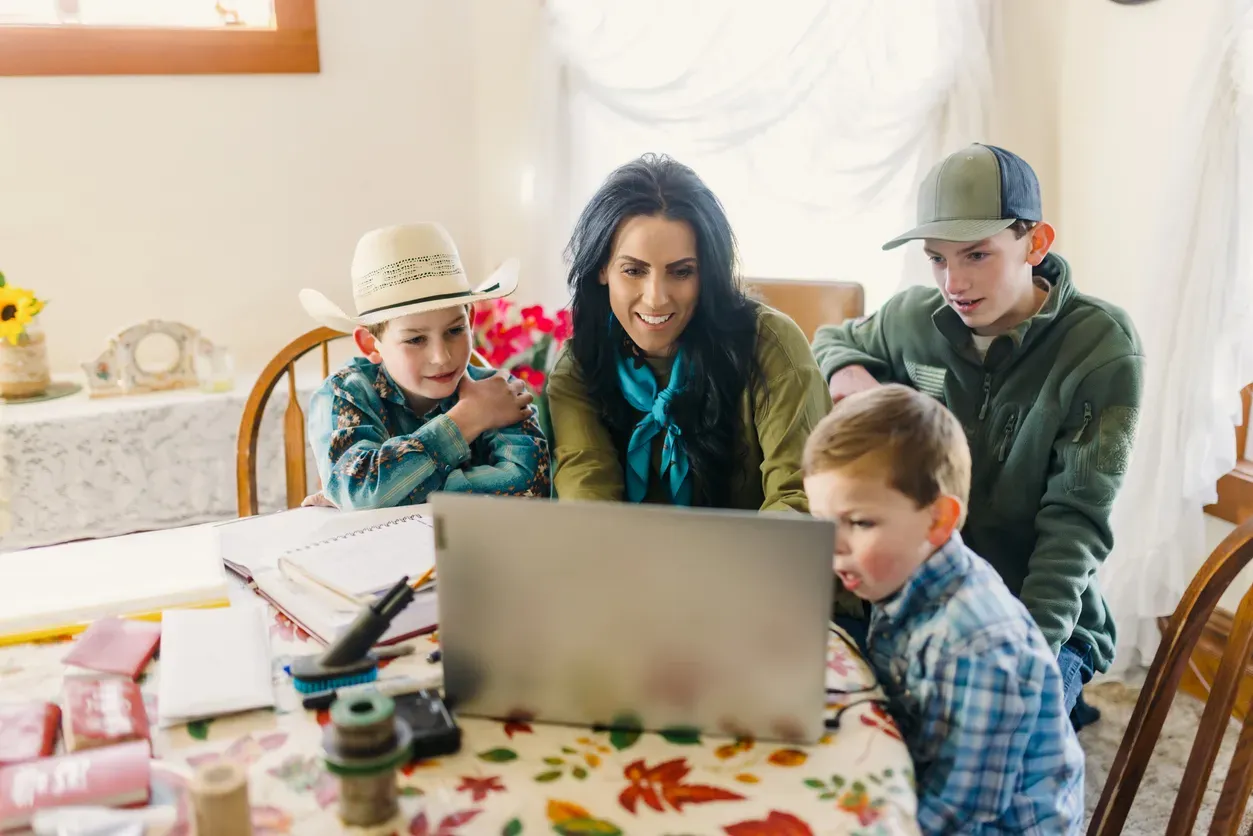 A smiling mother and three boys gather around a laptop at a dining table.