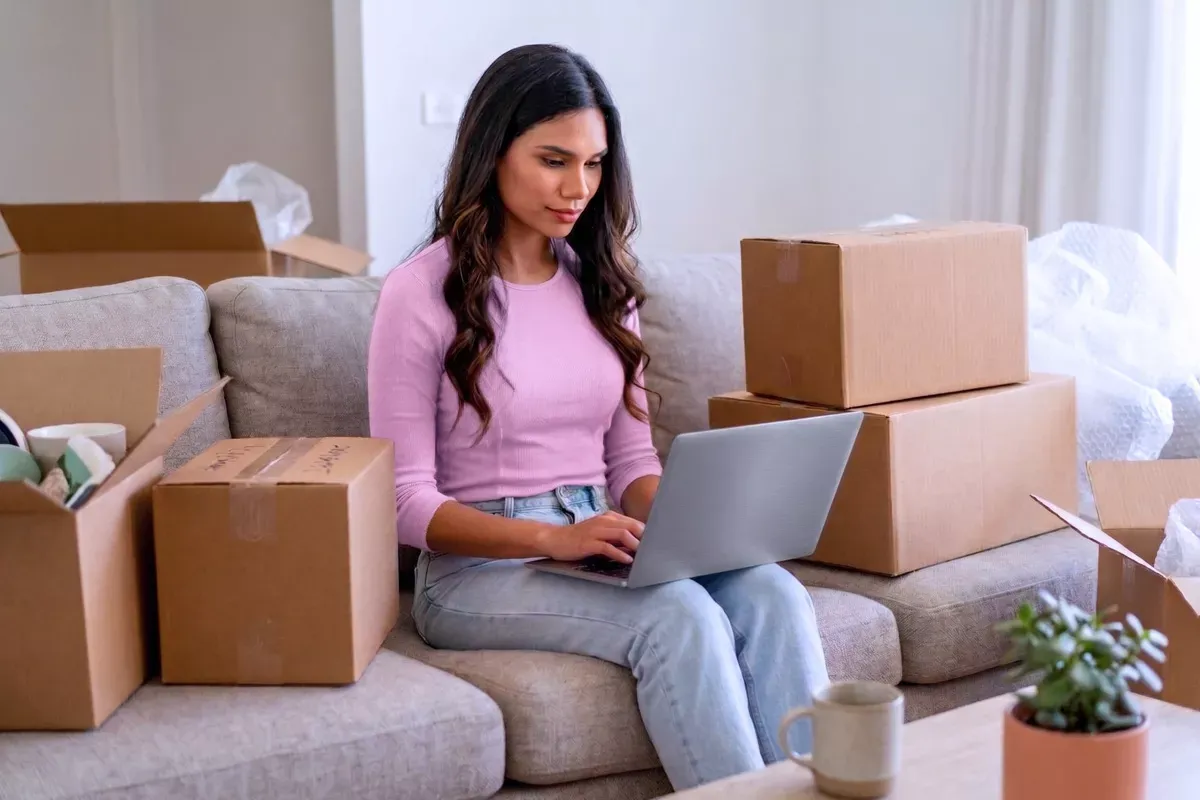 Woman in pink shirt using her laptop in between a bunch of moving boxes.