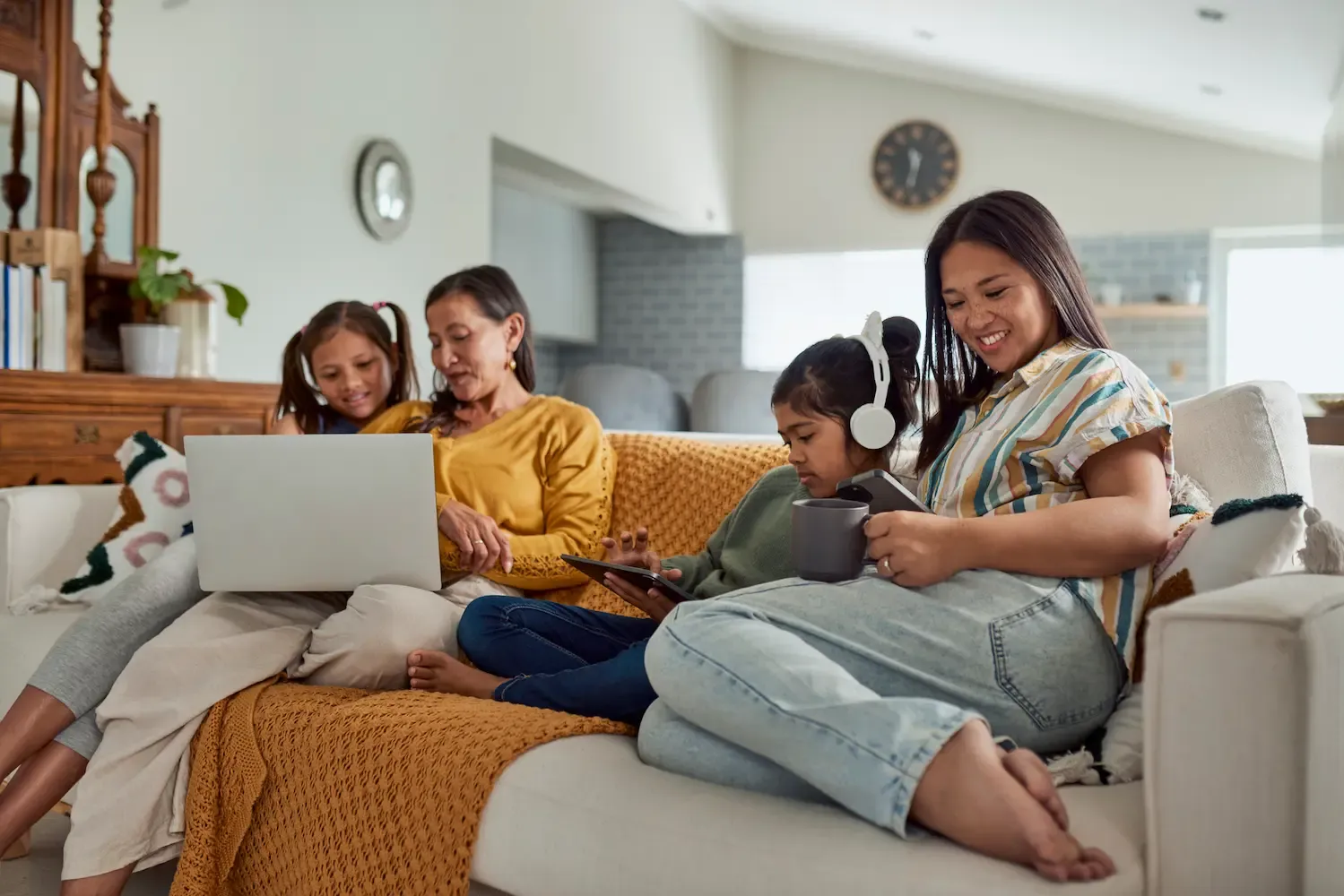 A multi-generational family of four sits together on a white sofa, each person engaged with their own digital device including a laptop, tablet, and smartphone.